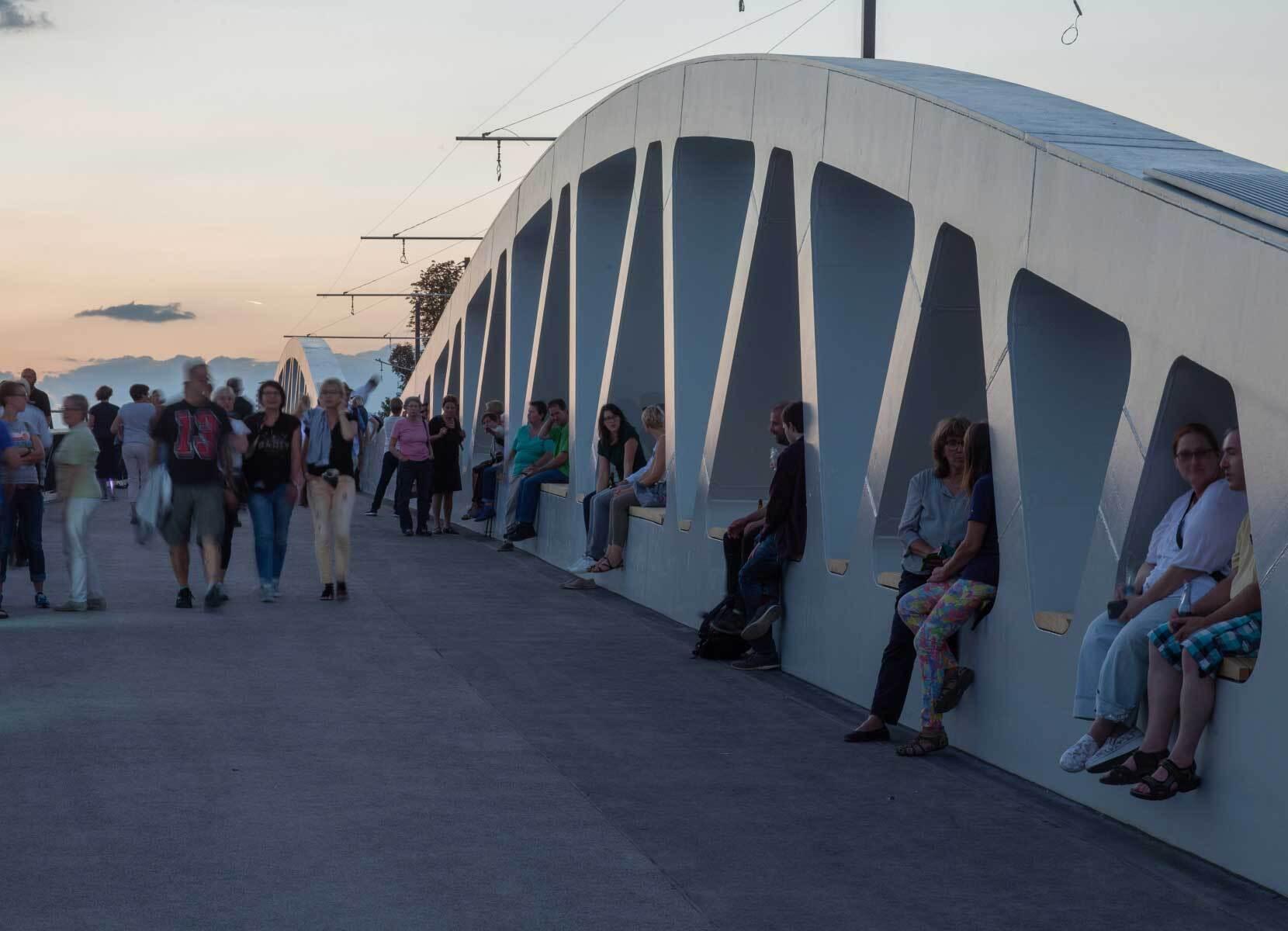 People walk over a modern bridge and sit in its chunky arches