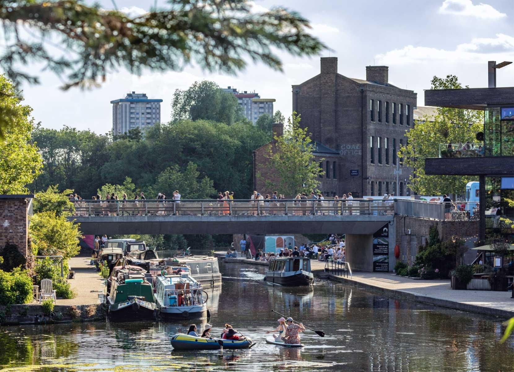 People walk over a bridge spanning a city canal, with narrow boats and inflatables in the water below.