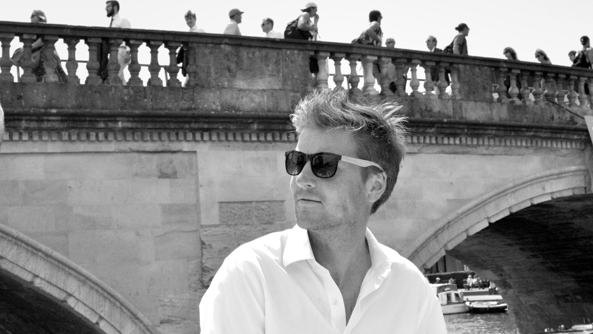 Black and white photo of Sam White wearing sunglasses in front of a classical stone bridge