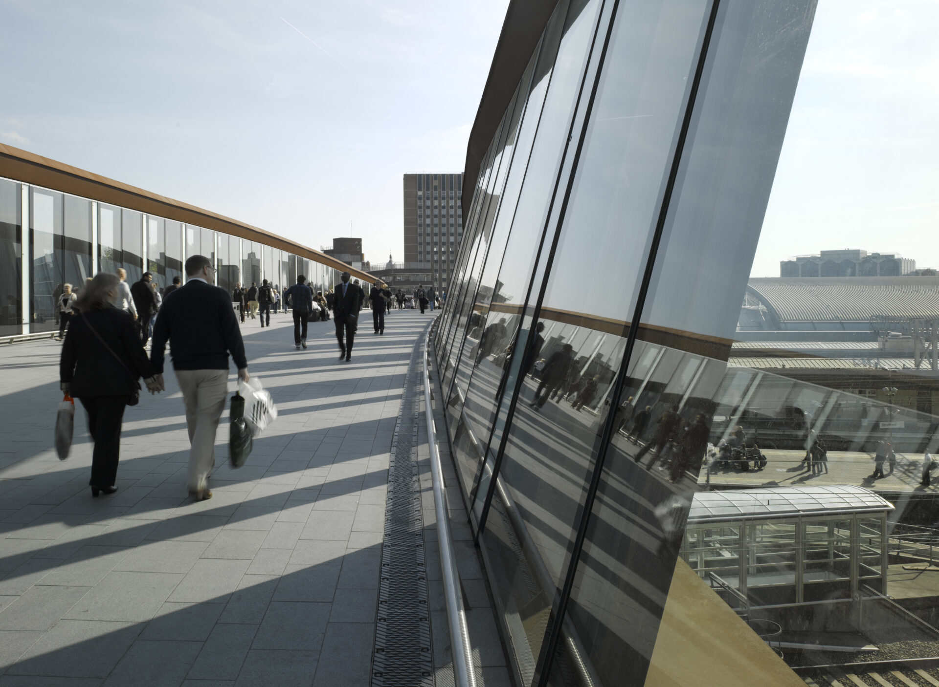 Reflection in the glass of the parapet of people walking across Town Centre Link Bridge at Westfield, Stratford